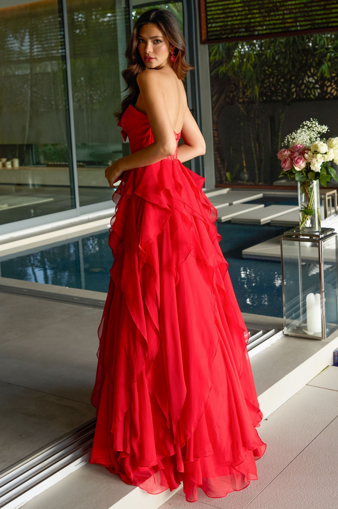 A woman wearing the Ava Presley 42209 Strapless Ruffled Prom Gown stands by an indoor pool, glancing over her shoulder. A glass vase with flowers rests on a table nearby.